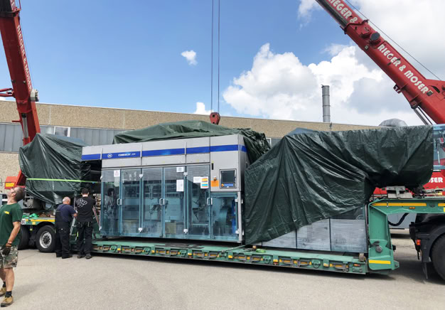 Große Maschine auf einem Lastwagen, teilweise mit grüner Plane abgedeckt, umgeben von mehreren Personen und zwei roten Kränen im Freien.