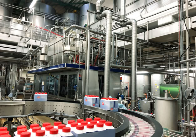 Produktionsanlage mit Förderband, das mit Kartons mit roten Verschlüssen beladen ist, umgeben von Rohren und Maschinen.